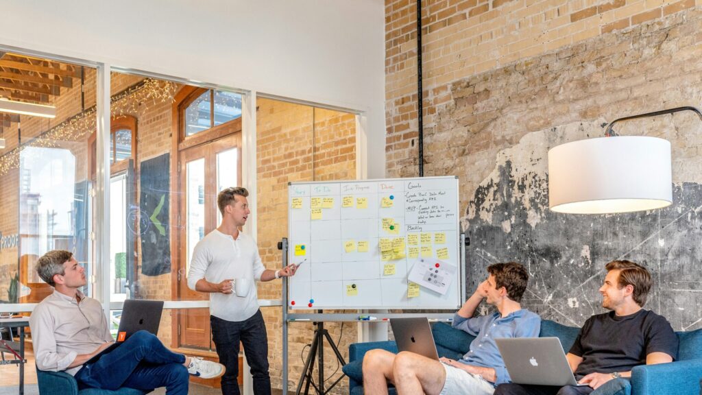 Four young man having a team meeting and one man is staying next to a white board explaining the tasks on the scrum board.